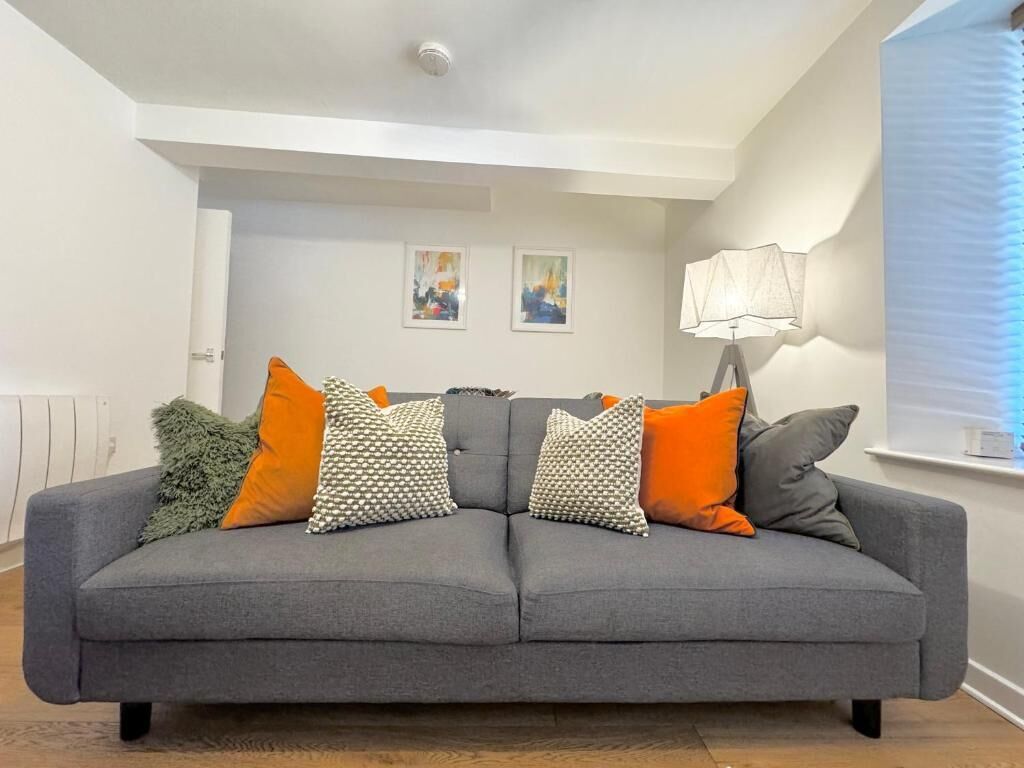 A grey sofa with multiple cushions, a wall-mounted lamp, and two framed artworks on the wall.