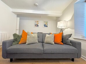 A grey sofa with multiple cushions, a wall-mounted lamp, and two framed artworks on the wall. - Nordic Suites (Ulverston)