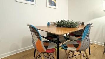 Un área de comedor con una mesa de madera y dos sillas con diseños coloridos, una planta en maceta sobre la mesa, y dos cuadros abstractos en la pared.