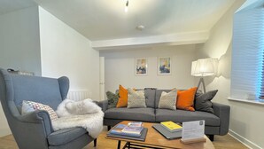 Un salon comprenant un fauteuil gris, un canapé gris avec des coussins à motifs, une table basse en bois avec des livres et un ordinateur portable, et une lampe.