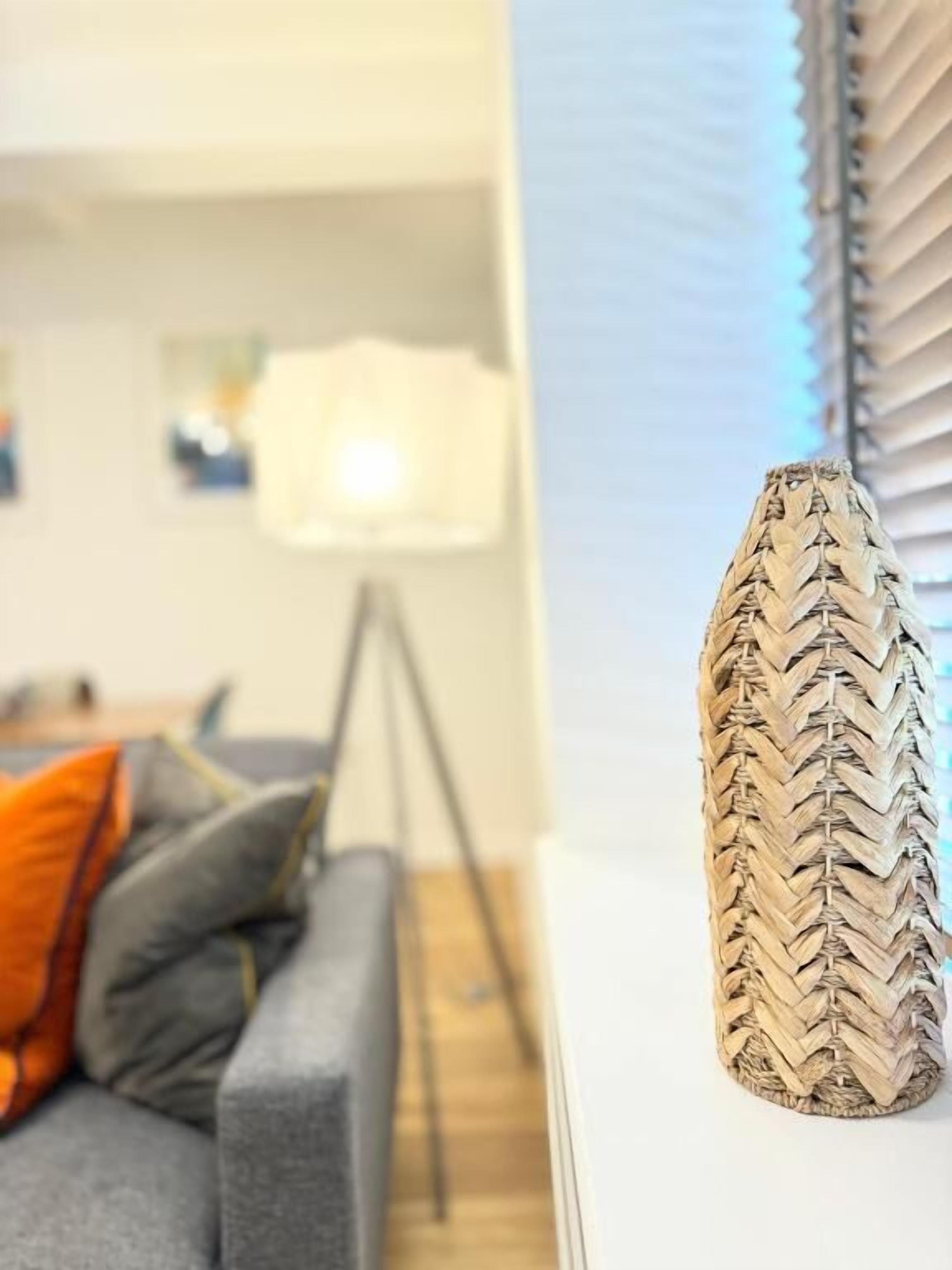 A room with a grey sofa, a wooden vase, and a window with blinds.