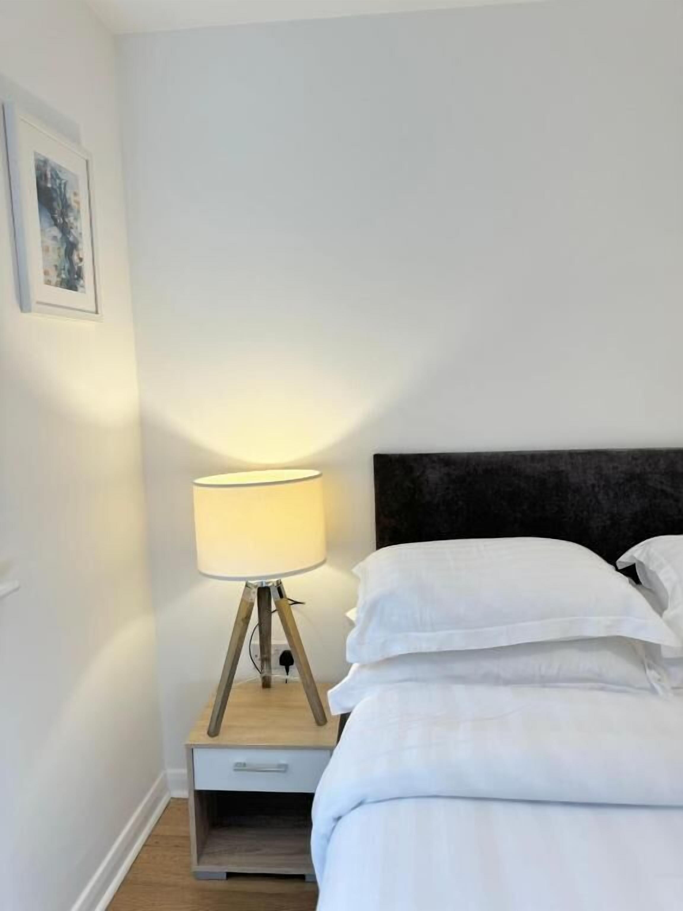 A neatly made bed with white linens, a bedside lamp on a wooden nightstand, and a framed picture on the wall.
