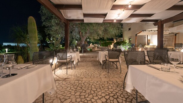 An outdoor dining area with tables and chairs, a stone floor, and a covered section with hanging lights. - Relais Torre Marabino (Ispica)
