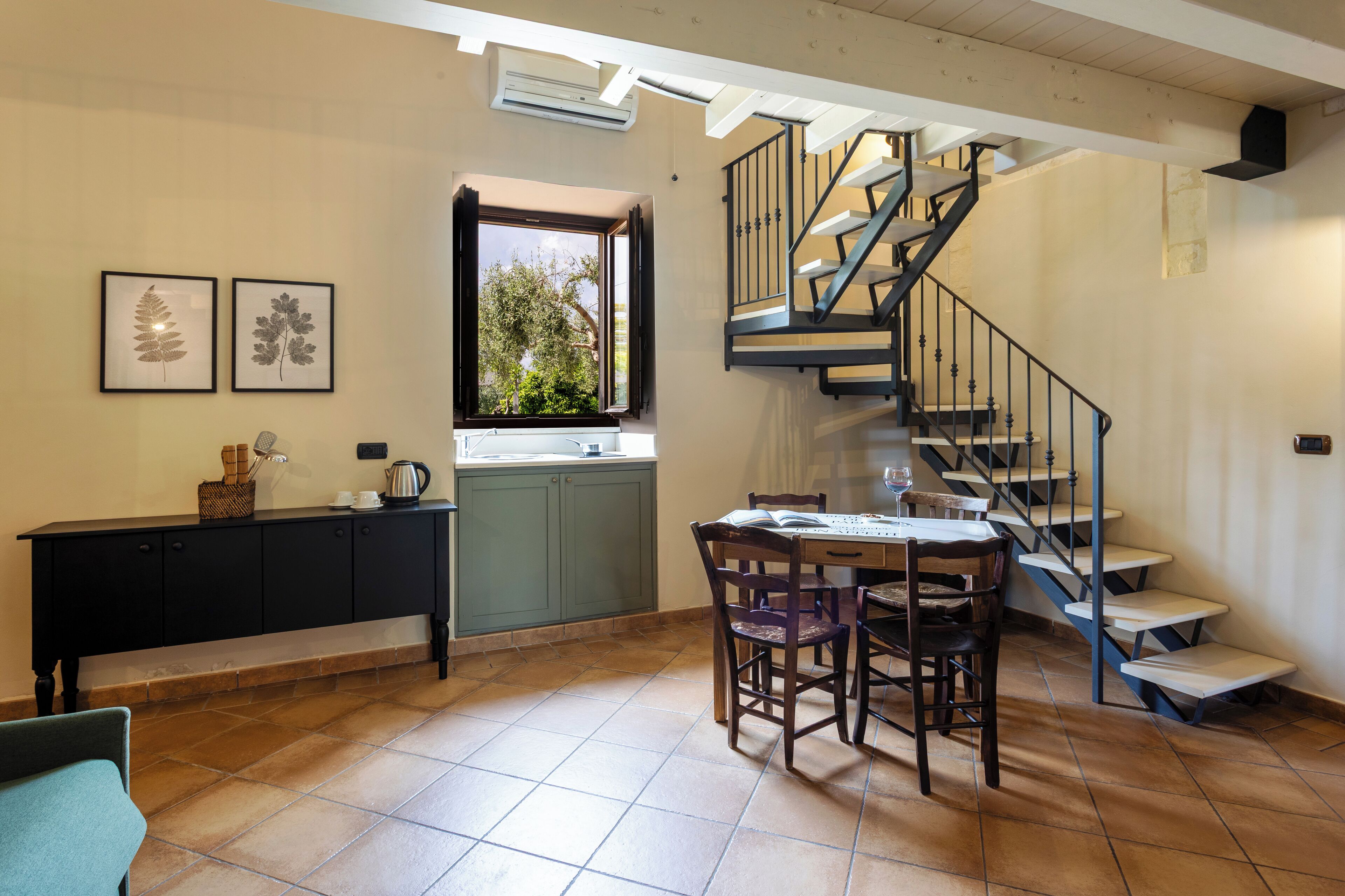 Una cocina con una mesa de comedor y sillas, una ventana con vista a follaje, una escalera y un armario negro con una cesta y una tetera encima.