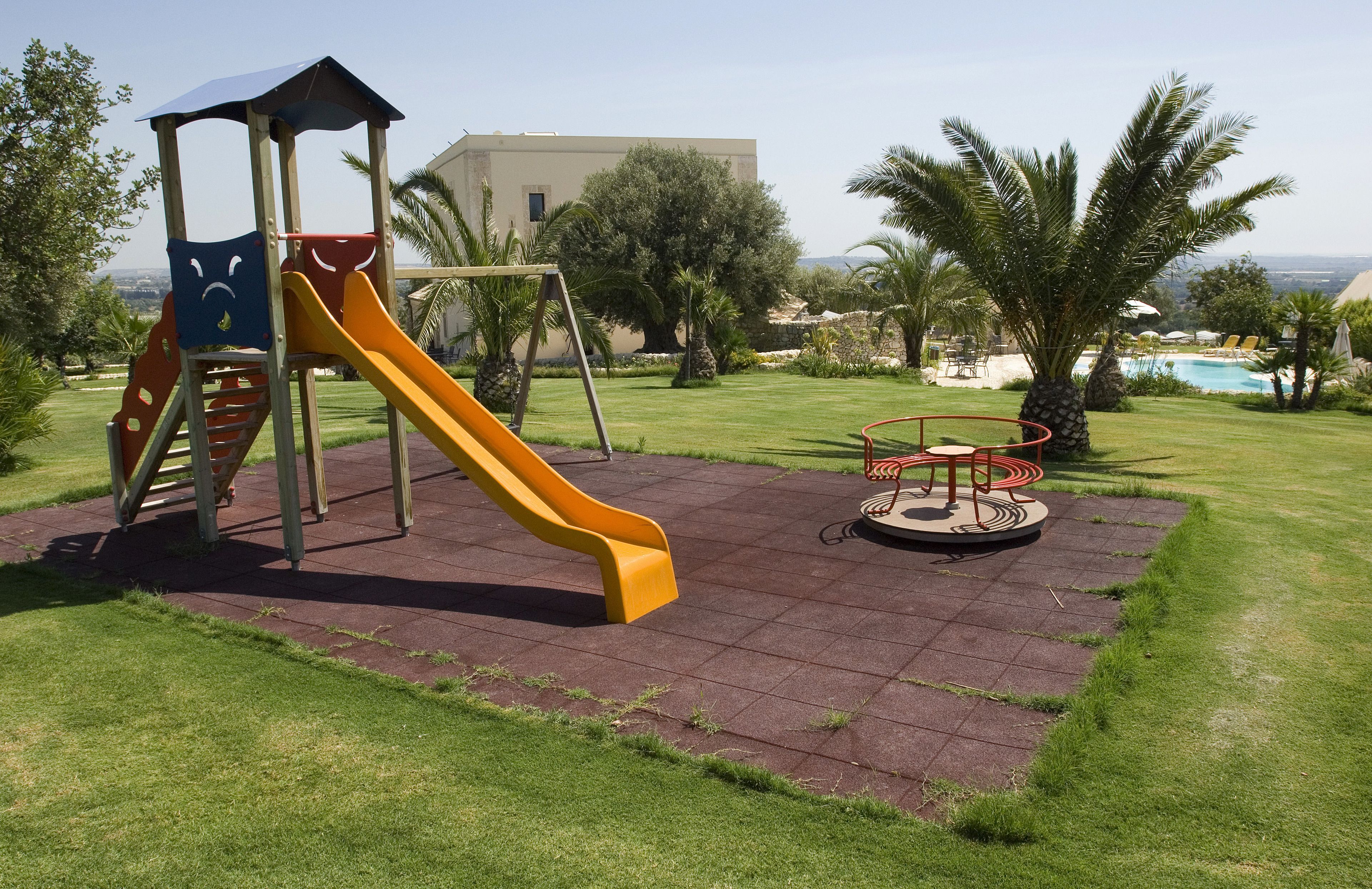 A playground with a slide and swings, surrounded by a well-maintained lawn and palm trees, with a pool in the background.