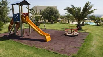 A playground with a slide and swings, surrounded by a well-maintained lawn and palm trees, with a pool in the background.