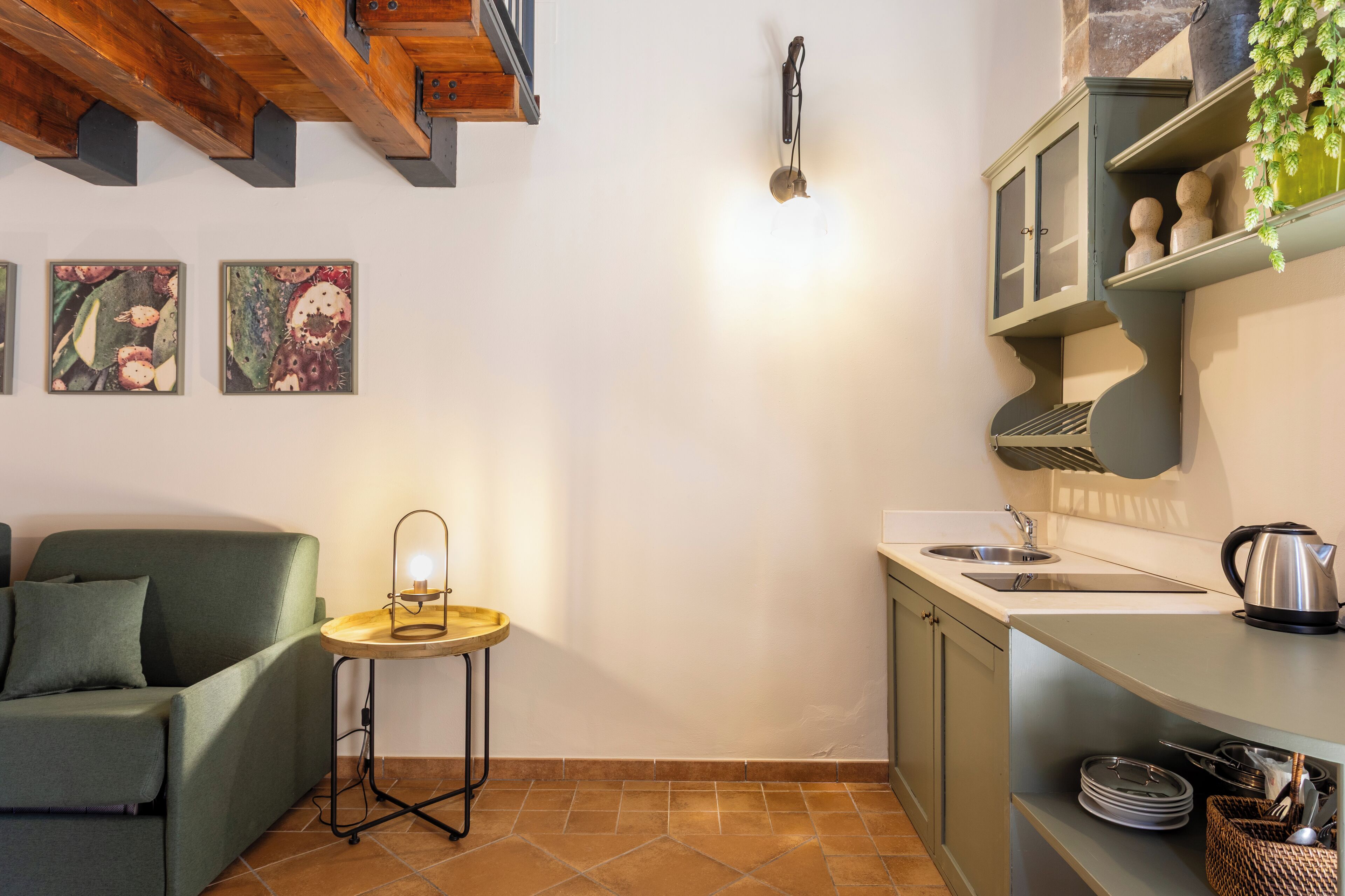 A compact kitchen with a sink, stove, and a small table with a light. There is a green sofa and a wall-mounted shelf with decorative items.