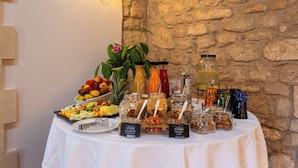 A buffet table with various food items and drinks, including fruits, salads, and jars of nuts and seeds, set against a stone wall. - Relais Torre Marabino (Ispica)