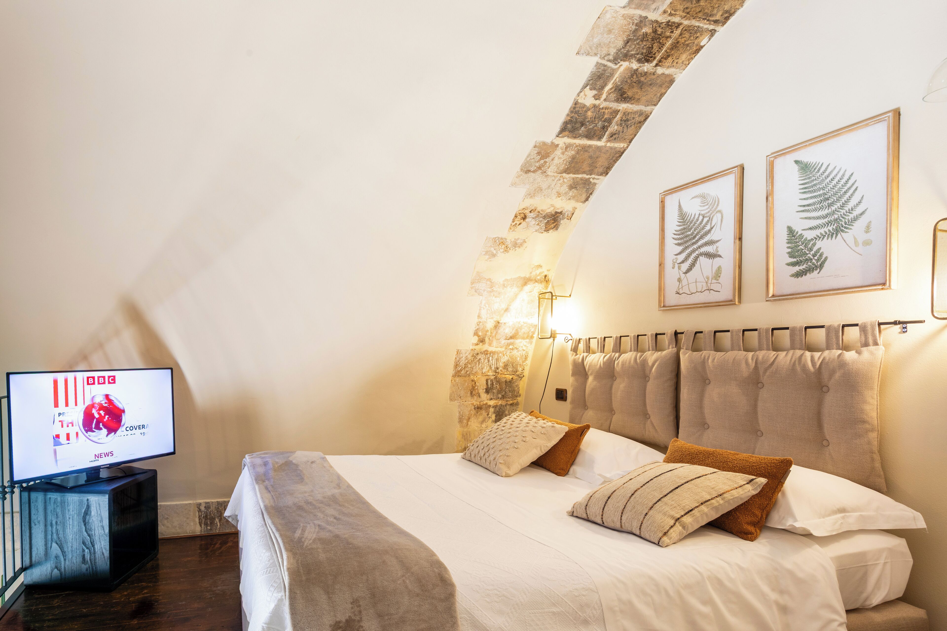 A bedroom with a stone wall, a bed with multiple pillows, a television on a stand, and framed botanical prints on the wall.