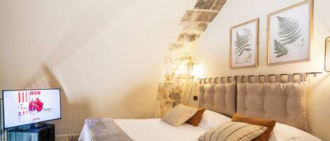 A bedroom with a stone wall, a bed with multiple pillows, a television on a stand, and framed botanical prints on the wall.