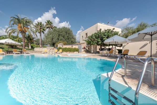 A swimming pool with clear blue water, surrounded by lounge chairs and umbrellas, with palm trees and a building in the background. - Relais Torre Marabino (Ispica)