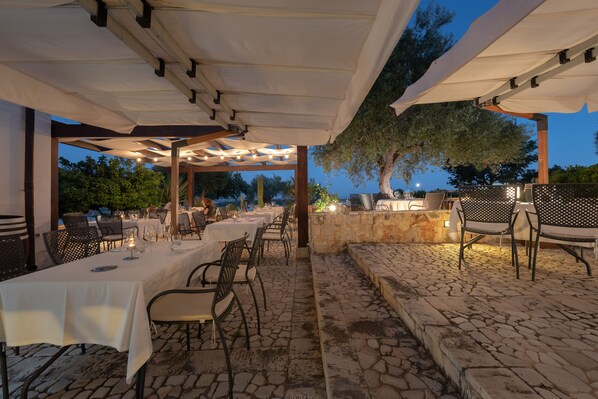 Un espacio exterior para comer con mesas y sillas, suelo de piedra y una estructura de pérgola.