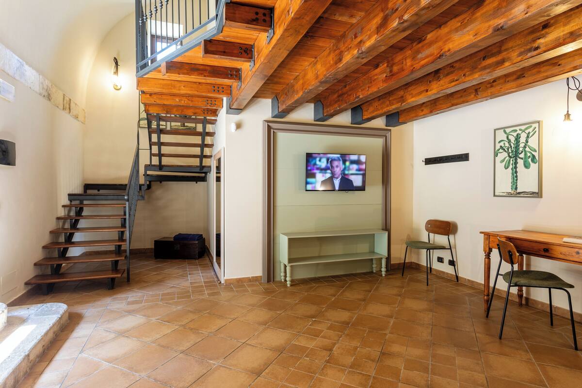 A spacious living area with a wooden staircase, a television, a bench, and a desk.