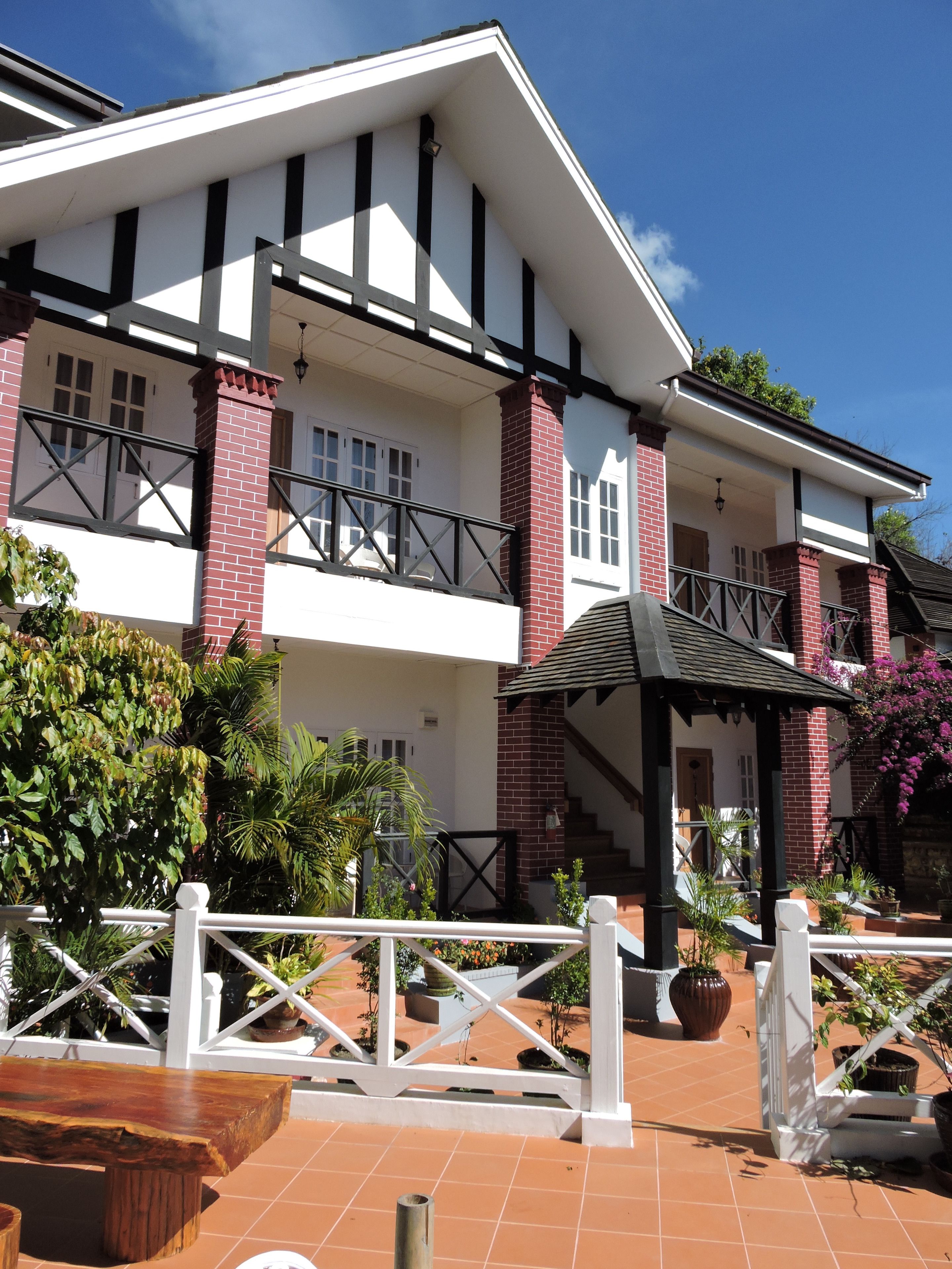 A two-story building with a white facade and red brick accents, featuring a balcony with black railings and a covered patio area with potted plants.