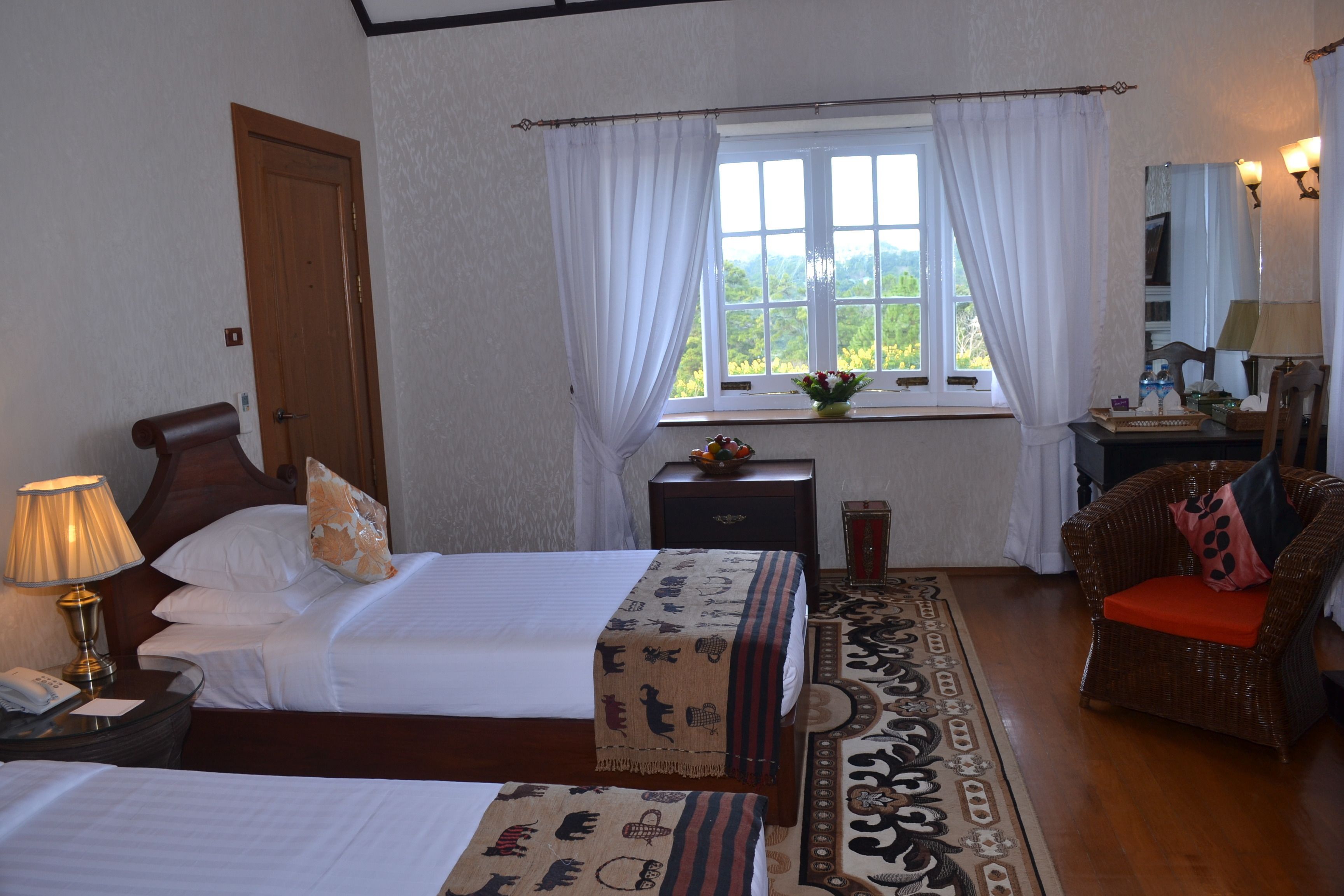 A hotel room with two beds, a wooden door, a window with white curtains, a nightstand with a lamp, a wicker chair, and a mirror with lights.