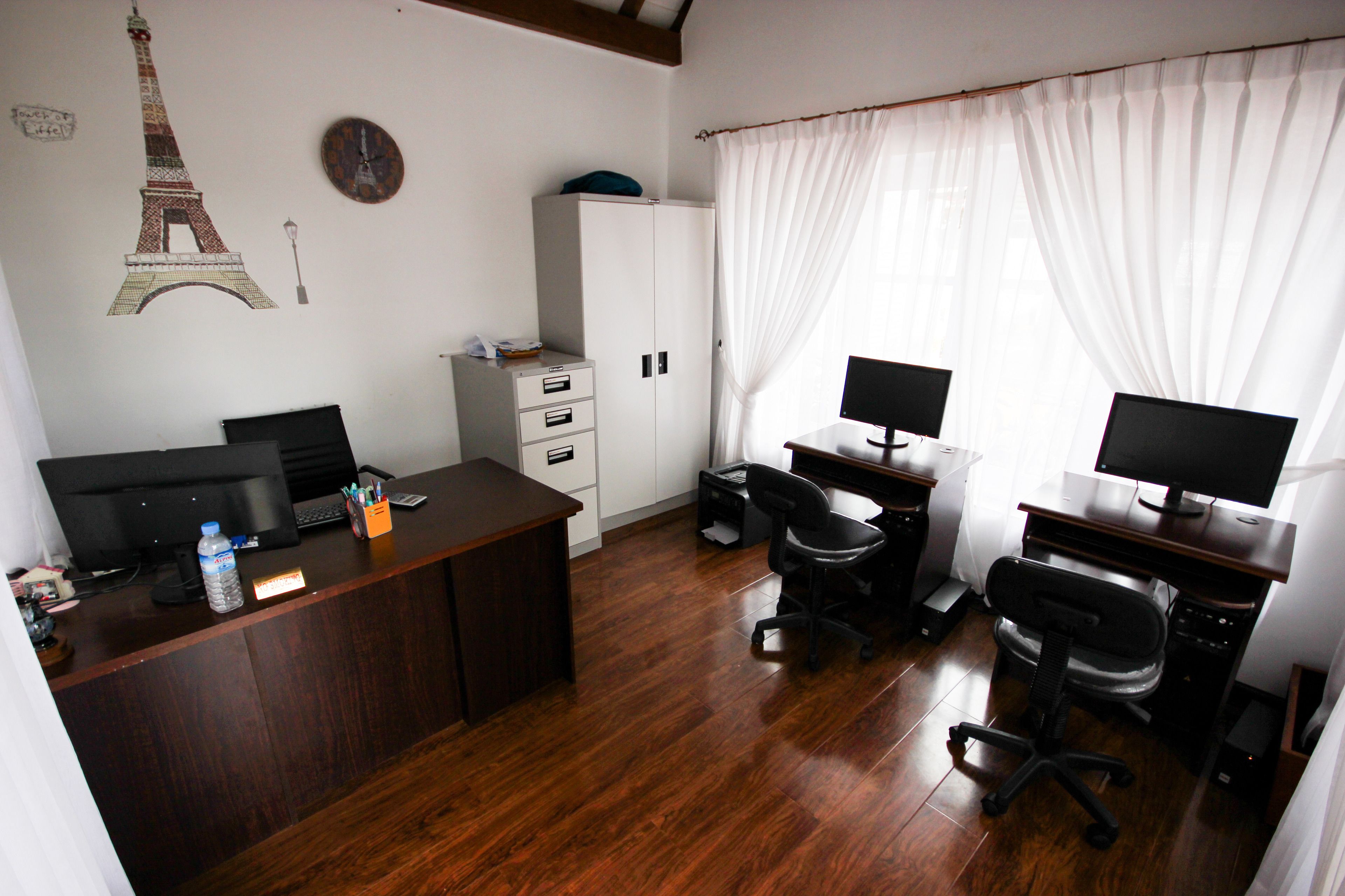 Une pièce avec un bureau en bois, deux écrans d’ordinateur, une chaise, une armoire et un mur orné d’une horloge et d’une représentation de la tour Eiffel.