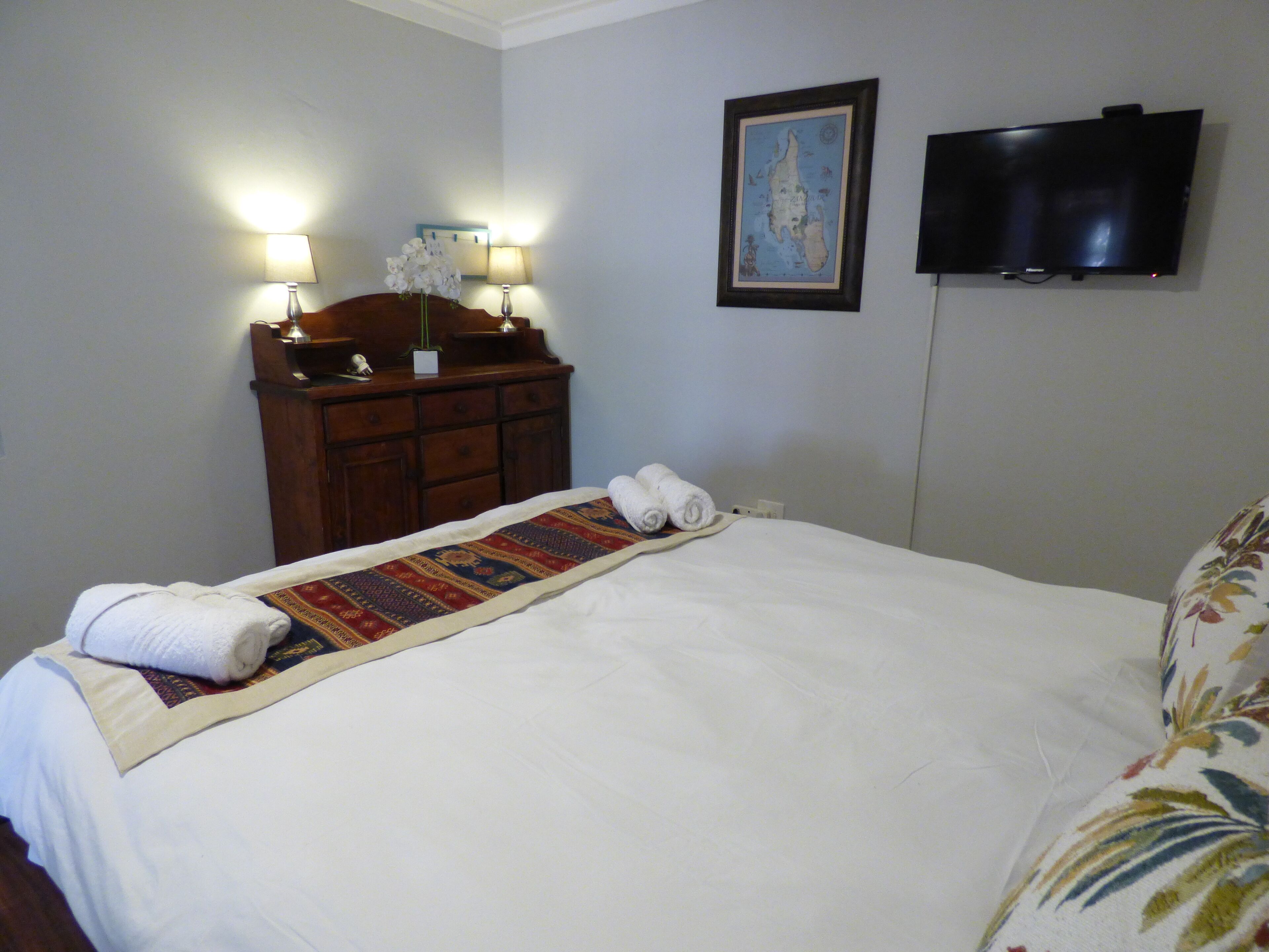 A neatly made bed with a patterned bedspread and rolled towels, a wooden dresser with lamps, and a flat-screen TV mounted on the wall.