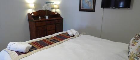 A neatly made bed with a patterned bedspread and rolled towels, a wooden dresser with lamps, and a flat-screen TV mounted on the wall.