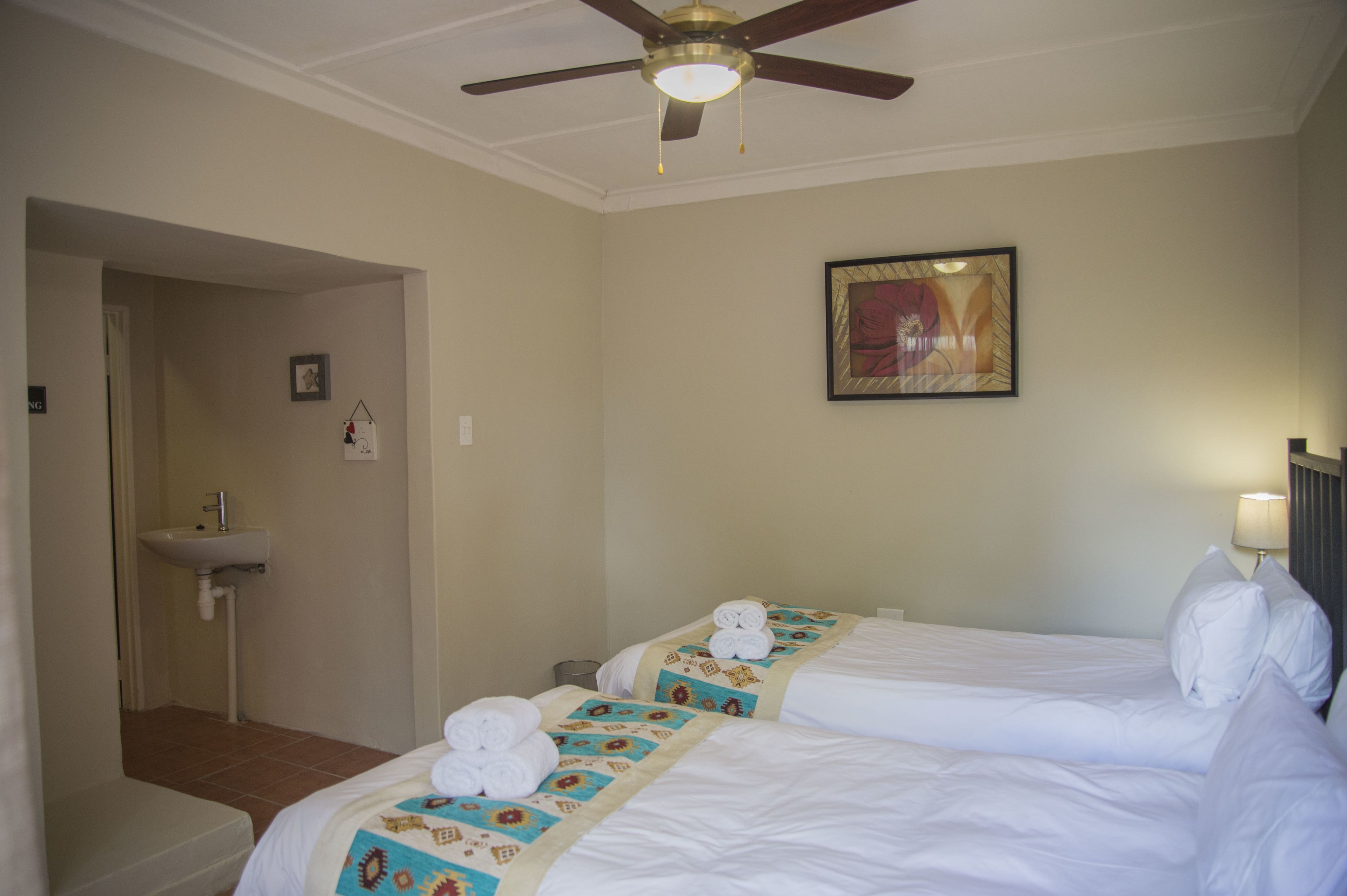 Habitación de hotel con dos camas, un ventilador de techo, un cuadro en la pared y un baño con lavamanos.