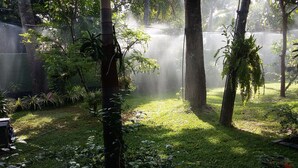 Een weelderige tuin met verschillende planten en bomen, een bewateringssysteem en een groene hek op de achtergrond.