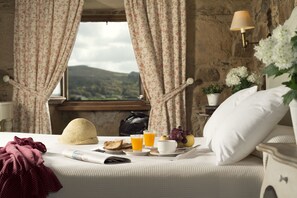 Una cama bien tendida con vistas al exterior, una lámpara de noche y una bandeja de desayuno con tostadas, jugo y fruta.