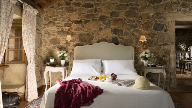 A stone-walled bedroom with a bed, two bedside tables, a window with curtains, and a small table with flowers and a camera. - A Casa da Torre Branca (Santiago de Compostela)