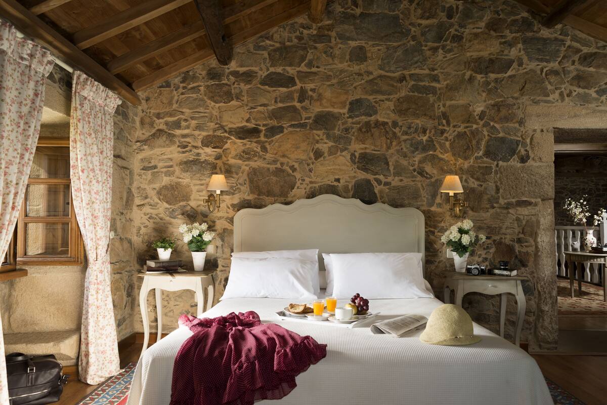 A stone-walled bedroom with a bed, two bedside tables, a window with curtains, and a small table with flowers and a camera.