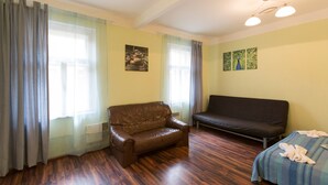 A room with a brown leather sofa, a black sofa, and a bed with a striped bedspread. There are two windows with curtains, and a painting on the wall. - Apartment for Friends (Prague)