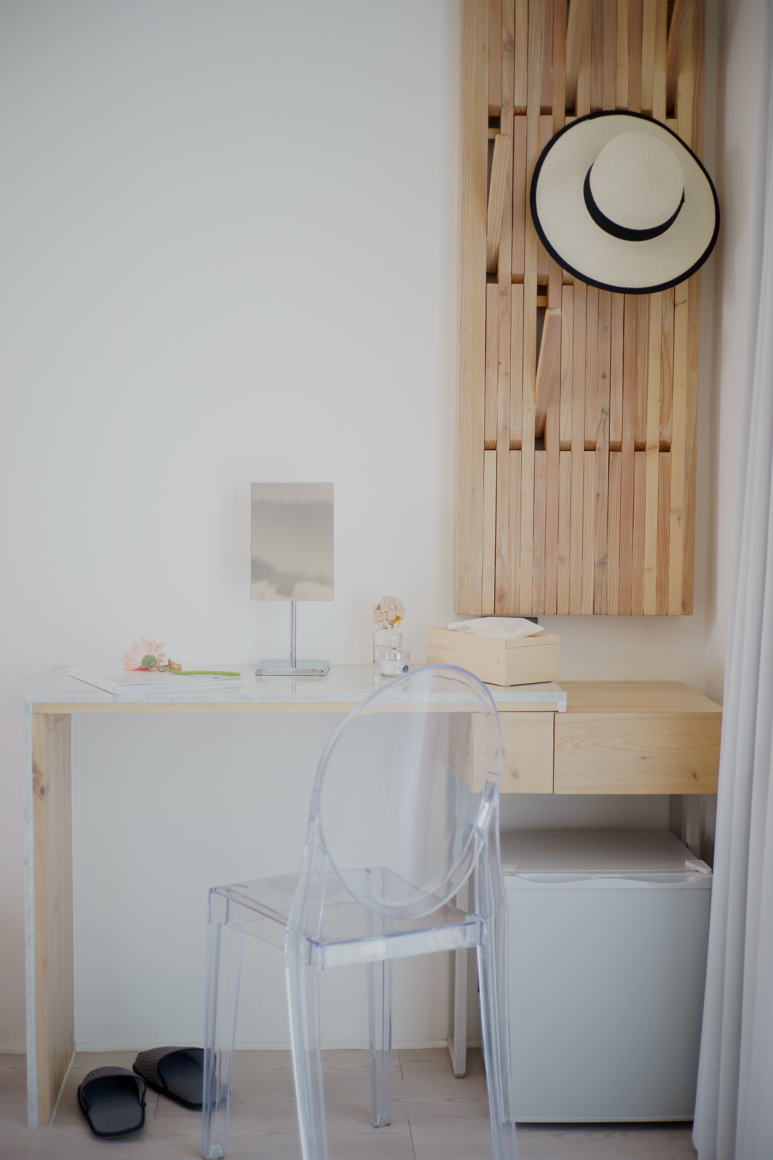 A minimalist room with a wooden desk, a clear plastic chair, a wooden cabinet, and a wall-mounted hat.