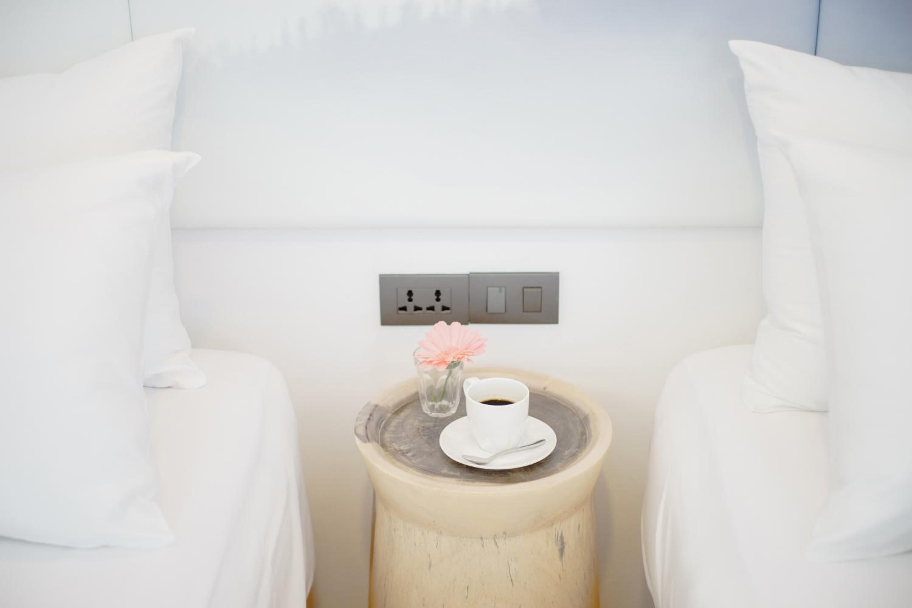 Een witte kamer met een nachtkastje waarop een kop koffie en een roze bloem staan.