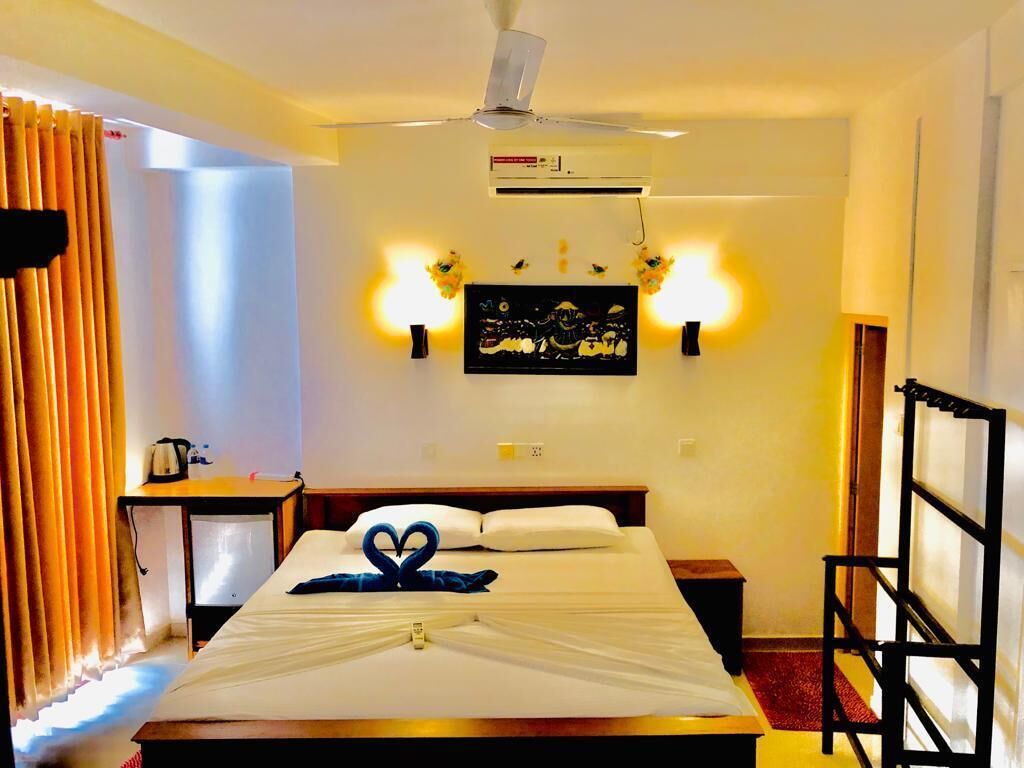Une chambre d’hôtel comprenant un lit, un ventilateur de plafond, un appareil de climatisation, une table de chevet, une lampe et une œuvre d’art accrochée au mur.