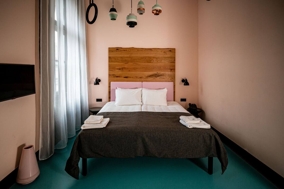 A neatly made bed with a wooden headboard, a bedside table with a lamp, and a window with sheer curtains.