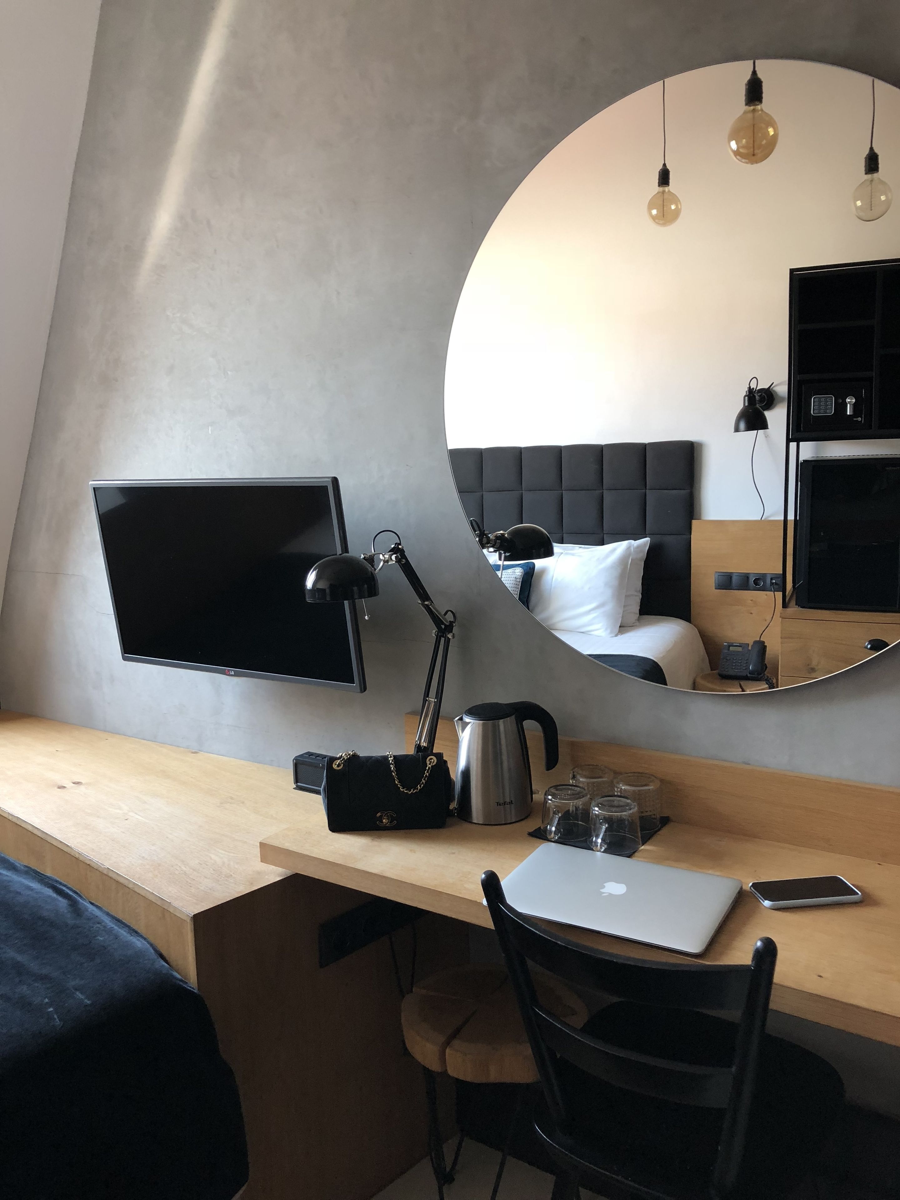 Ein modernes Hotelzimmer mit Schreibtisch aus Holz, einem Laptop, einem Smartphone, einer Kaffeemaschine, einer Handtasche, einem Spiegel, einem Bett mit schwarzem Kopfteil und einem Fernseher.