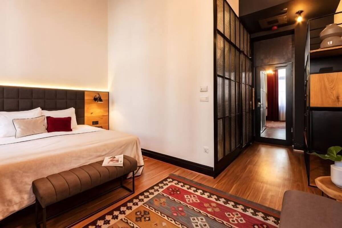 A hotel room with a bed, a bench, a patterned rug, a wooden floor, and a hallway leading to another room.