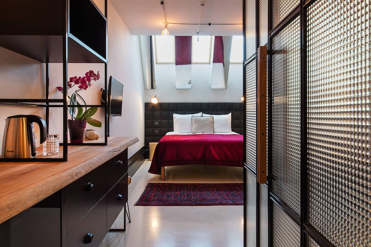 A modern hotel room with a wooden desk, a bed with a red bedspread, a large mirror, and a view of the city through the window.