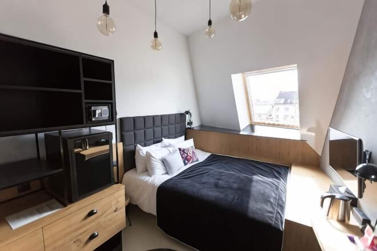 A bedroom with a bed, a desk, a chair, a TV, and a window.