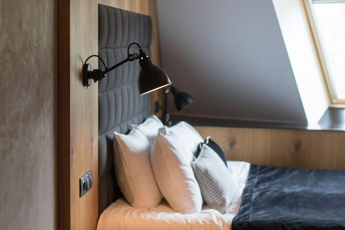 A bedroom with a wooden headboard, a bed with white and gray bedding, a bedside lamp, a telephone, and a skylight.
