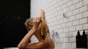 Una persona en una bañera con una pared de azulejos detrás. Hay dos botellas sobre el borde de la bañera.