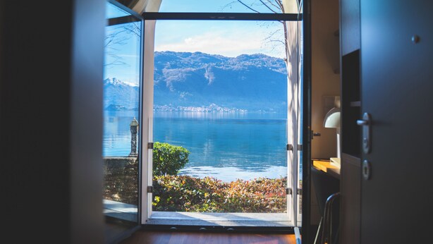 A room with a large arched window offering a view of a lake, mountains, and a distant cityscape.