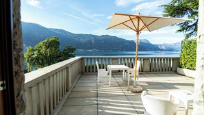 A balcony with a view of a lake and mountains, a white table and chair, and an umbrella.