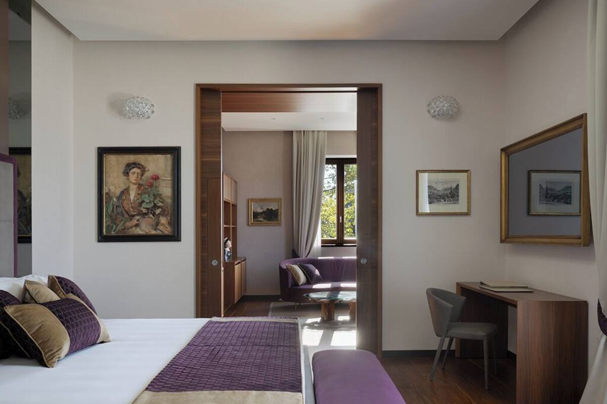 A hotel room with a large bed, a desk, and a chair. There is a door leading to another room.
