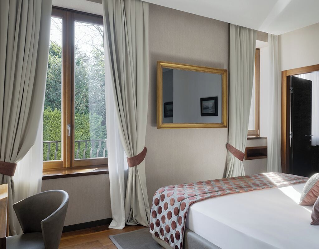 A hotel room with a bed, a chair, a window with curtains, and a mirror on the wall.