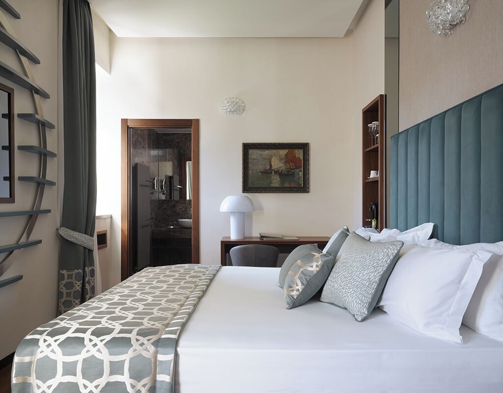 A hotel room with a bed, a desk, a chair, a lamp, a painting, and a wardrobe.