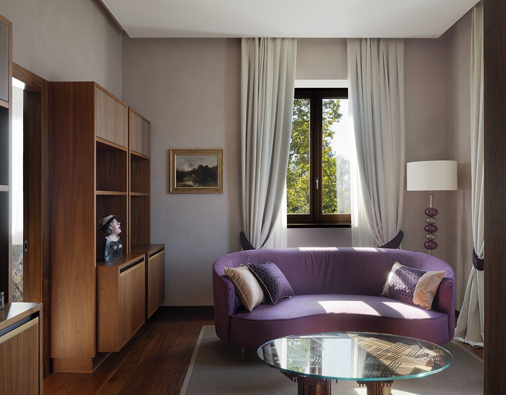 A modern living room with a purple sofa, a glass coffee table, wooden shelving, and a window with curtains.