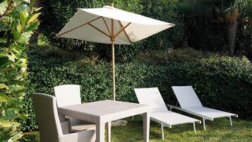 Un patio con una mesa, dos sillones, una sombrilla y abundante vegetación de fondo.