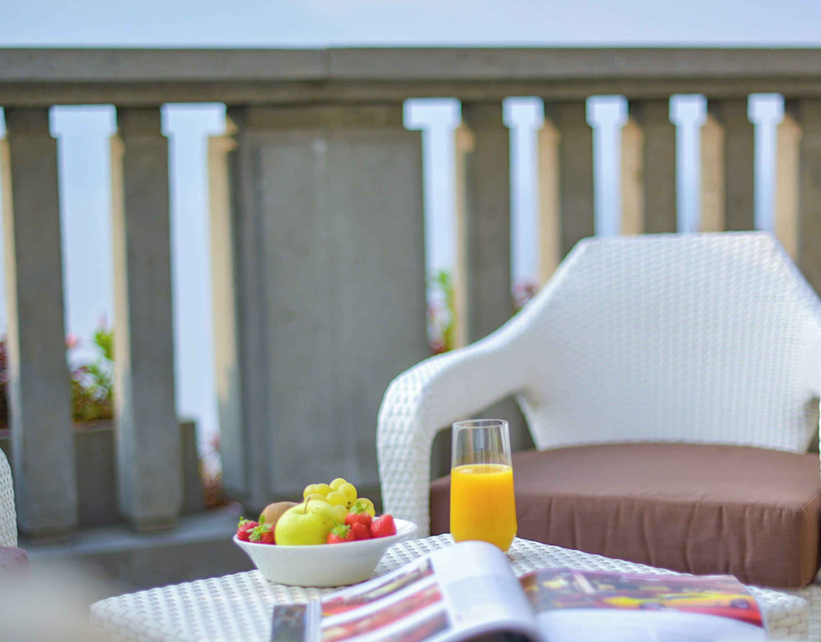 A dining area with a white wicker chair, a brown cushion, a bowl of fruit, and a glass of orange juice on a table.