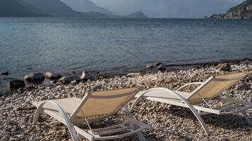 Dos tumbonas en una playa de guijarros con un lago y montañas al fondo.