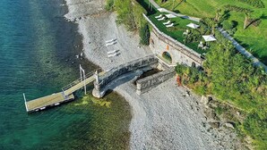 Ett flygbildsfoto av en fastighet vid kusten, med en stenbro som leder till ett grönområde med parasoller, en tennisbana och en strand med en stenstrand.