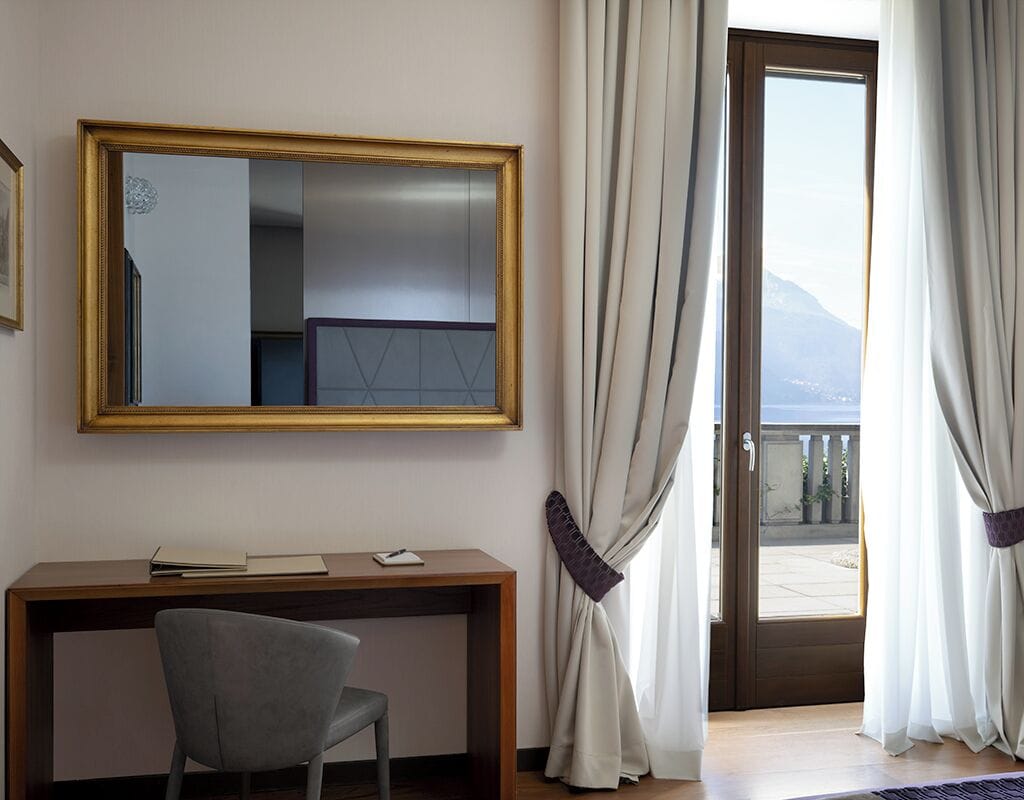 Une chambre d’hôtel équipée d’un bureau, d’une chaise et d’un grand miroir. La fenêtre donne sur un balcon avec des rideaux.