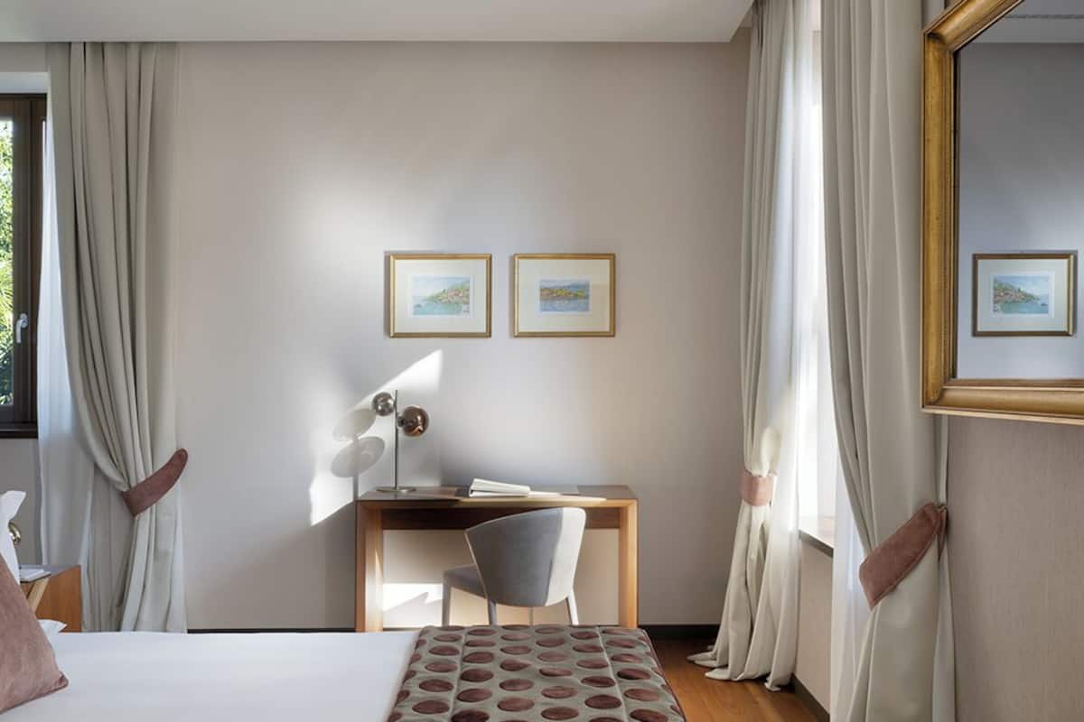 A hotel room with a bed, a desk, two framed pictures on the wall, and a window with curtains.